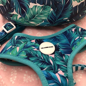 May include: A dog harness, collar, and bag set with a tropical leaf pattern in shades of green and blue on a pink background. The harness has a teal trim and a white circular patch that says "THE AUSSIE HOUND".