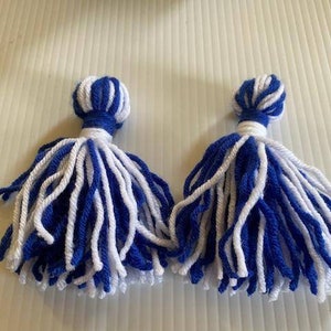 May include: Two blue and white yarn tassels. The tassels are made of white and blue yarn and are tied together at the top.