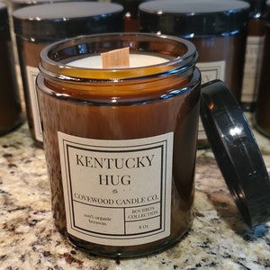 May include: A brown glass jar candle with a black lid, labeled "Kentucky Hug" from Covewood Candle Co. The label also states "Bourbon Collection" and "100% organic beeswax". The candle is 8 oz.