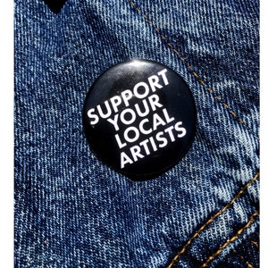 Op de afbeelding: Een zwarte knop met witte tekst die "Support Your Local Artists" zegt. De knop is vastgemaakt aan een blauw denim jack.