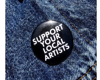 May include: A black button with white text that reads "Support Your Local Artists". The button is pinned to a blue denim jacket.