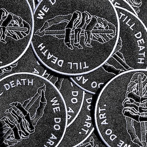 以下が含まれることがあります： 「We Do Art Till Death」というテキストが書かれた、黒と白の刺繍パッチがいくつか写っているクローズアップ。パッチには、さまざまな骨格の手のデザインが施されています。