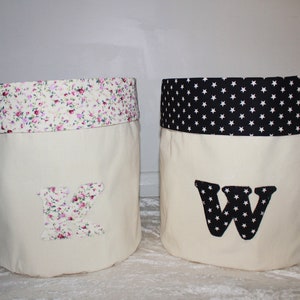 May include: Two fabric storage bins with white canvas sides and a contrasting fabric top. The bin on the left has a pink floral print top and a letter K on the front. The bin on the right has a black and white star print top and a letter W on the front.