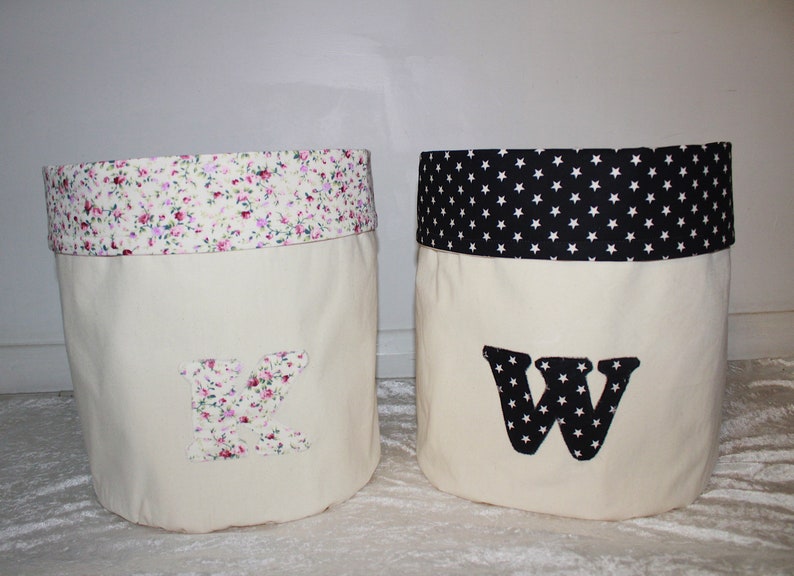 May include: Two fabric storage bins with white canvas sides and a contrasting fabric top. The bin on the left has a pink floral print top and a letter K on the front. The bin on the right has a black and white star print top and a letter W on the front.