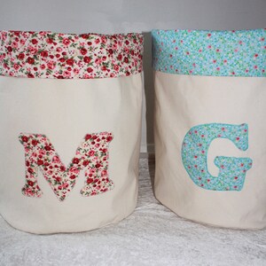 May include: Two fabric storage bins with floral patterns. The bin on the left has a red and white floral pattern and the letter "M" on the front. The bin on the right has a blue and white floral pattern and the letter "G" on the front.