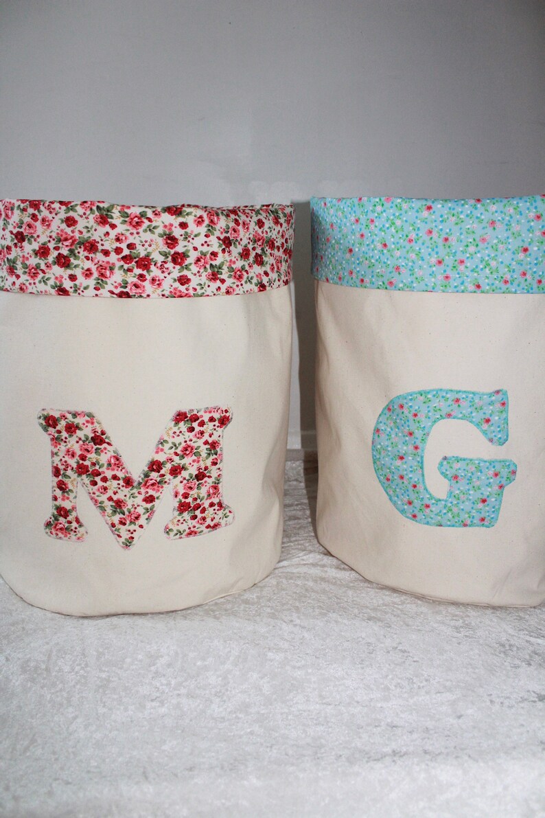May include: Two fabric storage bins with floral patterns. The bin on the left has a red and white floral pattern and the letter "M" on the front. The bin on the right has a blue and white floral pattern and the letter "G" on the front.