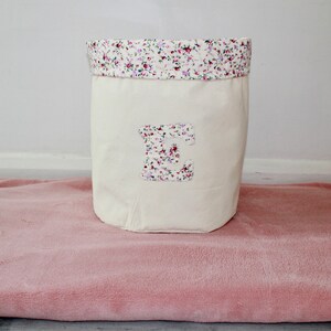 May include: A white canvas storage bin with a floral print top and a large letter "E" on the front. The bin is sitting on a pink fuzzy rug.