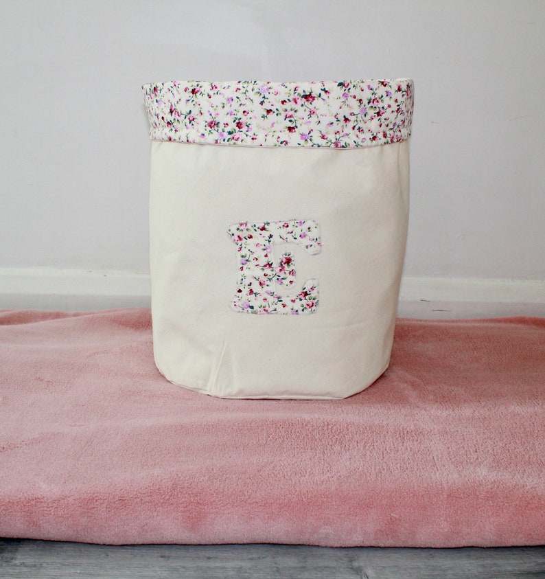 May include: A white canvas storage bin with a floral print top and a large letter "E" on the front. The bin is sitting on a pink fuzzy rug.