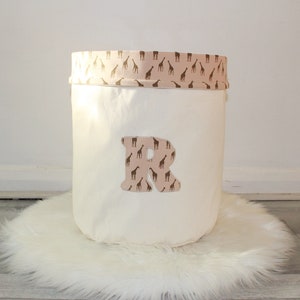 May include: White canvas storage bin with a pink and brown giraffe print trim. The bin has a large letter "R" on the front, also in the giraffe print. The bin is sitting on a white fluffy rug.