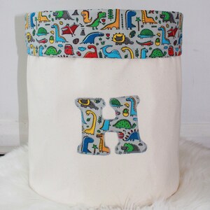 May include: A white canvas storage bin with a dinosaur print fabric top and a large fabric letter H on the front. The bin is sitting on a white fluffy rug.