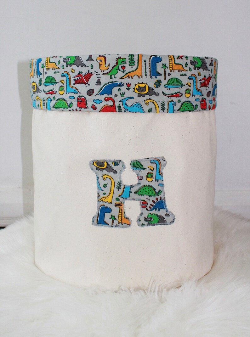 May include: A white canvas storage bin with a dinosaur print fabric top and a large fabric letter H on the front. The bin is sitting on a white fluffy rug.