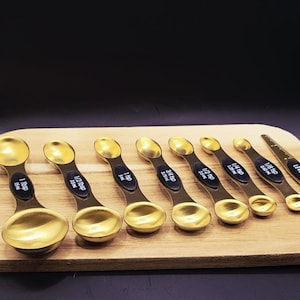 May include: A set of eight gold-colored measuring spoons with black text indicating measurements. The spoons are arranged on a wooden cutting board against a dark background. Measurements include 1 Tbsp, 1/2 Tbsp, 1 tsp, 3/4 tsp, 1/4 tsp, and 1/8 tsp.