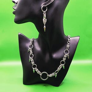 May include: A silver-toned chainmail necklace and a matching single earring featuring interconnected metal rings and small, pointed cone-shaped spikes. The necklace includes a larger central ring and several smaller spikes hanging from the chain links.