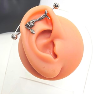 May include: A silver-toned industrial barbell piercing featuring a detailed snake design that wraps around the straight metal bar. The jewellery has a polished ball at each end and is displayed on a neutral-toned silicone ear model for demonstration.
