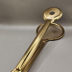 May include: A gold-toned metal kitchen tong featuring a circular, serrated-edge head on one side and a circular fine-mesh strainer on the other. The long, smooth handle connects the two ends, allowing the tool to open and close for gripping food items.