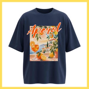 May include: A navy blue short-sleeve t-shirt featuring a rectangular graphic print. The design displays the text Aperol Spritz in orange and blue, with an illustration of a cocktail glass, fresh oranges, and a coastal Mediterranean villa scene.