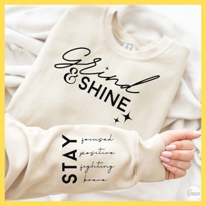 May include: A beige sweatshirt with the text "Grind & Shine" in black script on the front. The text "STAY" is printed vertically on the left sleeve, with the words "focused", "positive", "fighting", and "brave" printed below it.