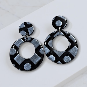 May include: A pair of black and silver geometric earrings. Each earring features a small, round stud and a larger, circular drop. The drop has a repeating pattern of silver shapes on a black background. The earrings are designed for pierced ears.
