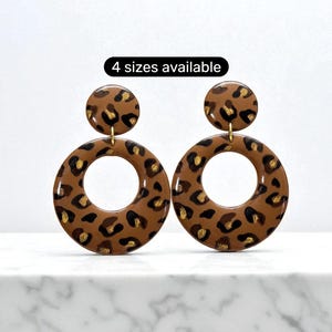 May include: A pair of brown and gold leopard print earrings. The earrings have a circular design with a smaller circle at the top. The text "4 sizes available" is in a black oval above the earrings.