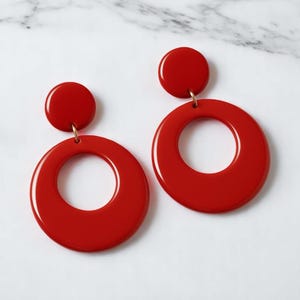 May include: A pair of bright red, retro-style earrings. Each earring features a small, circular stud connected to a larger, circular hoop. The earrings are made of a glossy material and hang from gold-colored hooks.