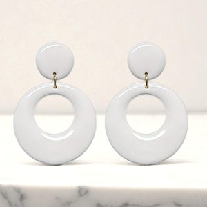 May include: A pair of white, retro-style earrings. Each earring features a small, circular stud connected to a larger, hollow circle. The earrings are suspended by a gold-colored metal hook.