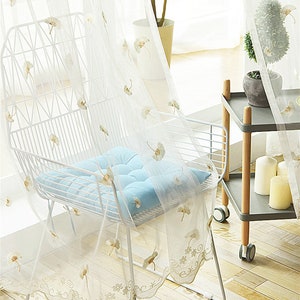 Può includere: Tende bianche e trasparenti con un delicato motivo floreale e un bordo in pizzo. Una sedia in metallo bianco con un cuscino blu chiaro è in primo piano.