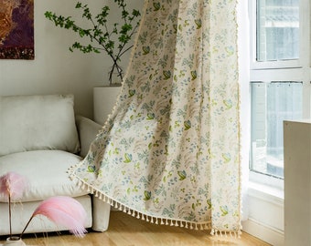 Puede incluir: Una cortina blanca y transparente con un patrón floral en tonos verdes y azules. La cortina tiene un ribete de borlas y está colgada en una ventana.