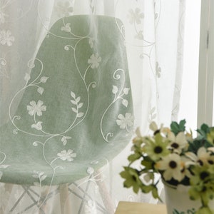Puede incluir: Cortinas blancas y transparentes con diseños florales bordados. Las cortinas están colgadas en una ventana y cubren parcialmente una silla verde.