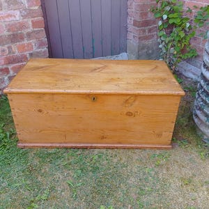 May include: A rectangular wooden storage chest with a natural light brown finish, featuring a prominent wood grain pattern, a flat top lid, and a small central metal keyhole escutcheon on the front panel, resting on a base with a moulded trim.