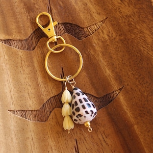 May include: A gold-colored keychain with a lobster clasp and a large ring. The keychain features a seashell pendant with black spots and three small, cream-colored flower-shaped beads. The background is a wooden surface.