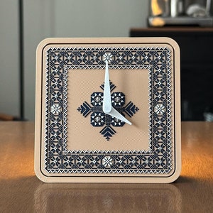 May include: A square clock with a beige background and a black and white geometric design. The clock has white hands and a black and white geometric design around the edge.