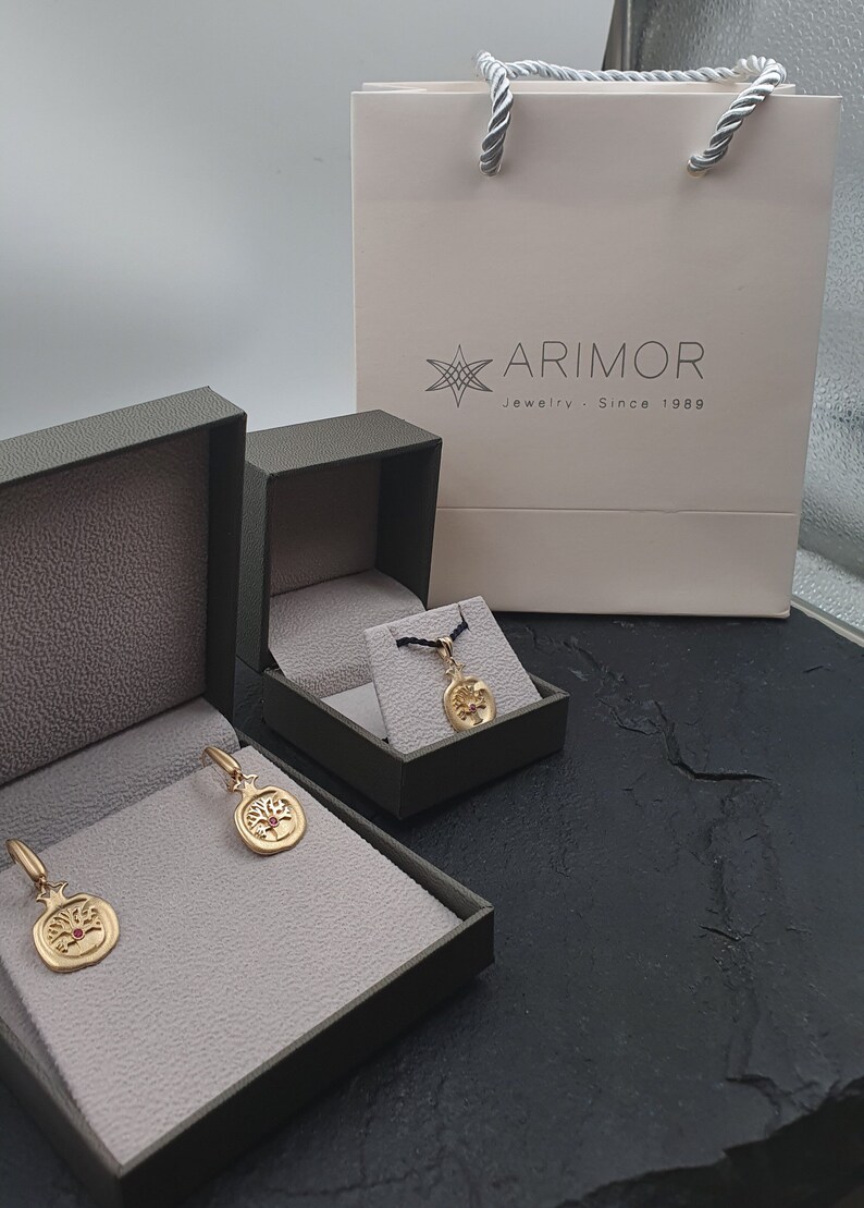 May include: Three green jewelry boxes with gold pomegranate earrings and a gold pendant necklace. The boxes are on a black surface. The pendant necklace is in a smaller box. The earrings are in a larger box. The boxes have a white label with the text "ARIMOR Jewelry - Since 1989".