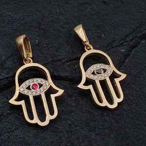 May include: Two gold hamsa hand charms with a detailed eye design. One charm has a red gemstone in the eye, and the other has a black gemstone.