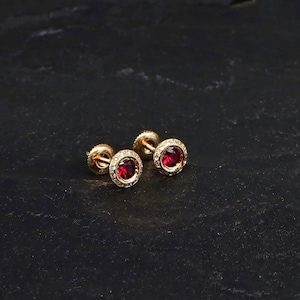 Puede incluir: Un par de pendientes de oro con una pequeña piedra preciosa roja en el centro de cada pendiente. Los pendientes están rodeados por un halo de piedras preciosas blancas más pequeñas.
