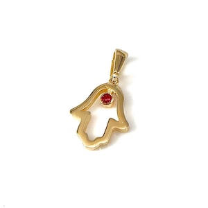 Puede incluir: Colgante de mano de Hamsa de oro con una piedra preciosa roja en el centro.