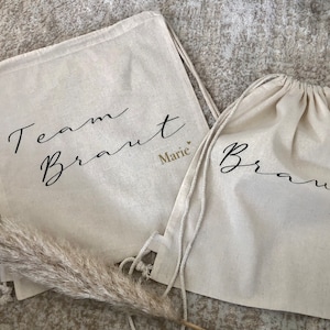 Pode incluir: Duas sacolas brancas de cordão com o texto "Team Braut Marie" e "Braut Luisa" escrito em caligrafia preta.