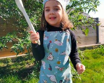Può includere: Un bambino con cappello e grembiule da chef, che tiene un cucchiaio di legno. Il grembiule è azzurro con un motivo a dinosauri. Il cappello ha una fascia blu abbinata. Il bambino sorride ed è all'aperto.