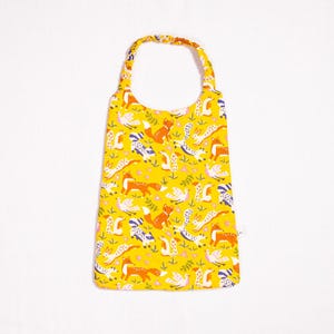 May include: A yellow bib with a woodland animal print featuring foxes, squirrels, birds, and flowers. The bib has a neck strap and a back closure.