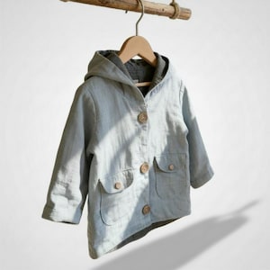 Puede incluir: Chaqueta azul claro con capucha y botones de madera. La chaqueta tiene dos bolsillos delanteros y está colgada de una percha de madera. La tela es suave y texturizada.