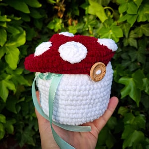 Op de afbeelding: Een gehaakte doos in de vorm van een paddenstoel met een rood fluwelen bovendeel en witte gehaakte stippen. De doos heeft een witte gehaakte basis en een houten knoop met de tekst "Liefde met" erop. Een lichtgroen lint is om de doos gebonden.