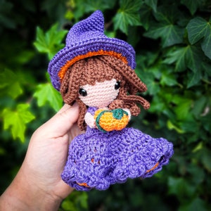 Op de afbeelding: Een paarse gehaakte heksenpop met een paarse en oranje hoed en een kleine oranje pompoen in haar hand.