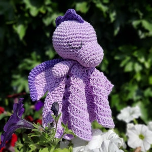 May include: A handmade crochet dragon in shades of purple. The dragon has a rounded head, small wings, and a textured body. The dragon is sitting among flowers, with a blurred green background.
