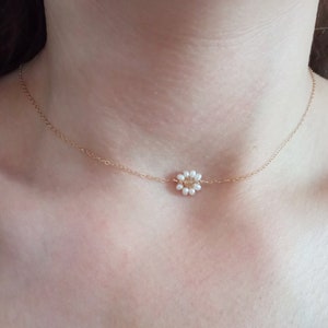 Pode incluir: Um delicado colar dourado com um pingente de margarida. O pingente é composto por pequenas pérolas brancas que circundam uma conta central dourada. O colar é exibido no pescoço de uma pessoa.