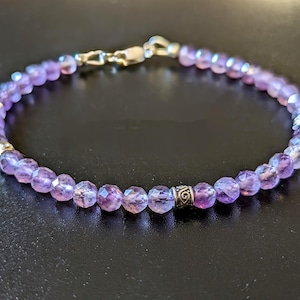 Pode incluir: Um bracelete de ametista roxa com detalhes prateados. O bracelete é feito de pequenas contas facetadas e tem um fecho prateado.