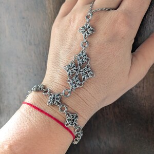 May include: A silver-toned chainmail hand flower bracelet featuring intricate interlocking metal rings arranged in geometric star-like patterns, connecting a wrist band to a single finger loop with a delicate chain.