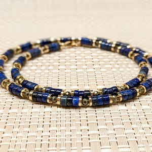May include: A double-strand necklace made of blue lapis lazuli and gold beads. The necklace is on a beige textured surface.
