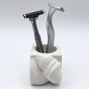 Puede incluir: Portaobjetos de baño de cerámica blanca con forma de figura humana con dos maquinillas de afeitar plateadas en su interior.