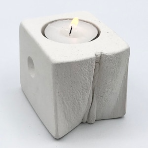 Puede incluir: Portavelas de hormigón blanco con una superficie texturizada y un pequeño agujero en el lateral. Una vela de té blanca está ardiendo dentro del portavelas.