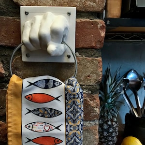 Könnte beinhalten: Ein weißer, handförmiger Keramik-Handtuchhalter an einer Ziegelmauer, der einen Metallring hält. Ein dekoratives Küchentuch mit Fischillustrationen in Orange, Blau und Schwarz hängt an dem Ring. Das Handtuch hat einen gelben Rand und einen gemusterten blauen Abschnitt.