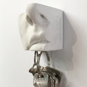 May include: White ceramic key holder shaped like a mouth with a nose and lips. A set of keys is hanging from the bottom of the holder.